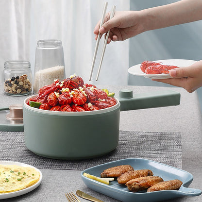 A sage green electric multi-cooker with a handle is filled with red spicy crayfish and garnished with garlic. A person's hands are shown using white chopsticks to lift food from the pot, with plates of food and glass jars arranged on a gray table runner.