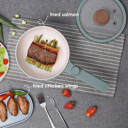 Fried salmon on a plate with a frying pan, chicken wings, and vegetables on a gray tablecloth.