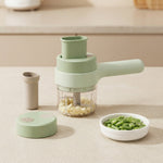 A light green manual food chopper with a clear container filled with chopped garlic, shown on a beige countertop with its lid, a small accessory cup, and a white bowl of green beans.